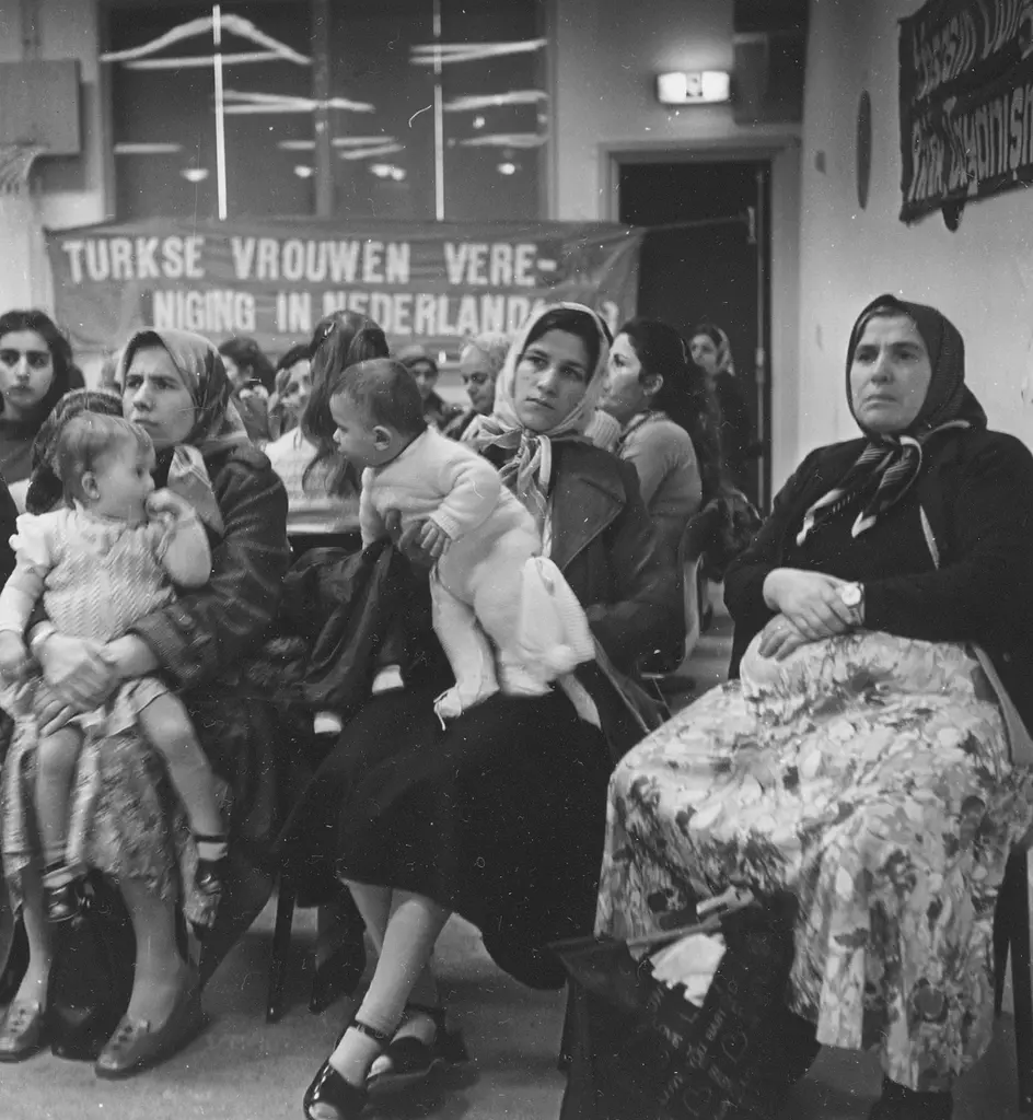 Group of women with some small children sitting on chairs with a canvas with Turkish Women's Association in the Netherlands in the background