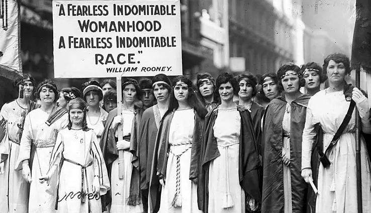 Groep vrouwen in witte lange jurken met een protestbord waarop staat: A Fearless Indomitable Womanhood - A Fearless Indomitable Race - William Rooney