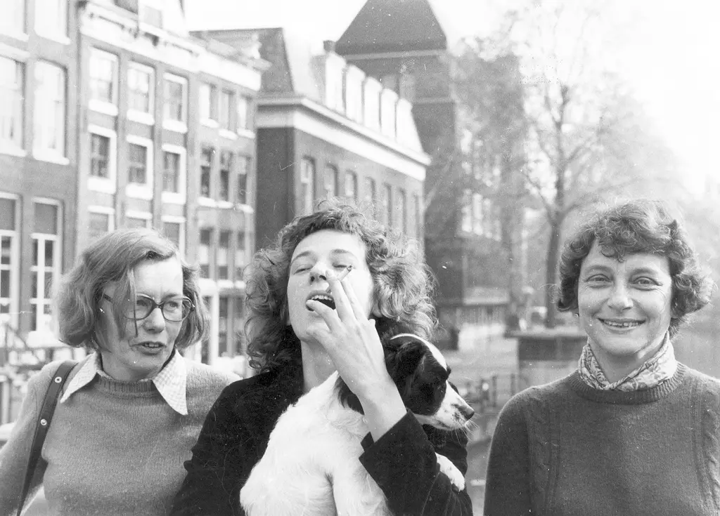 Drie vrouwen naast elkaar poserend voor de grachten van Amsterdam. Zwart-wit foto. Middelste vrouw rookt een sigaret. 