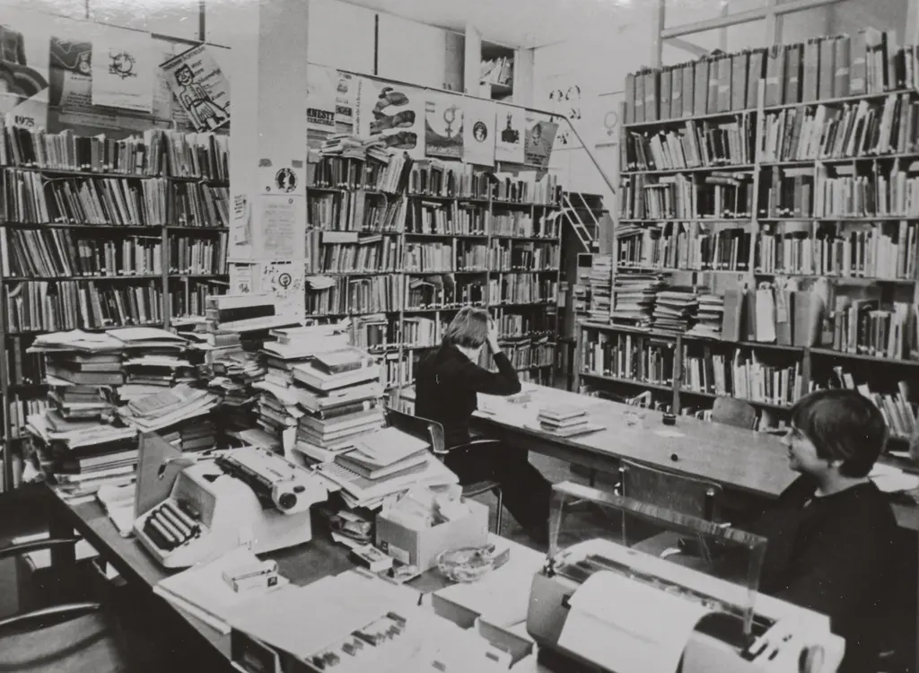 2 medewerkers aan het werk aan tafels met hoge stapels boeken erop en omringd door volle boekenkasten