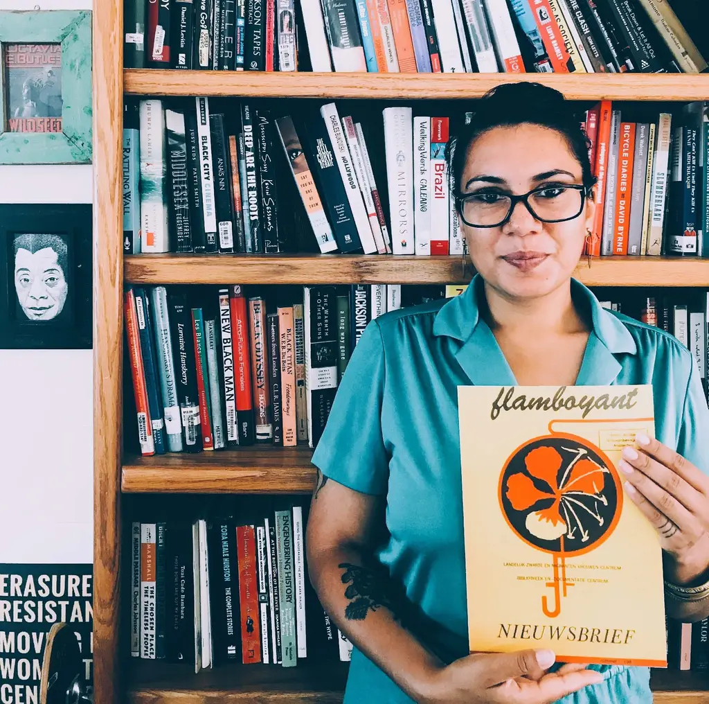 Vrouw met zwarte bril en blauwe jurk staat voor boekenkast met tijdschrift Flamboyant in haar handen