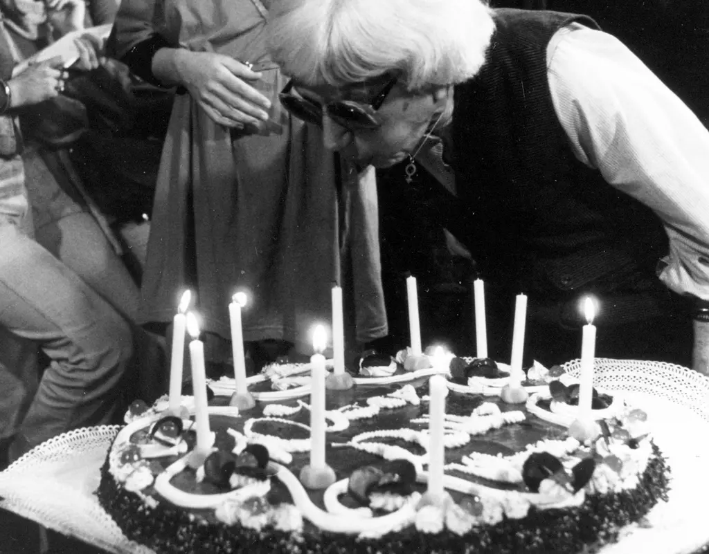 Woman with glasses blows out candles from a large cake. Around her, people watch her blow out the candles.