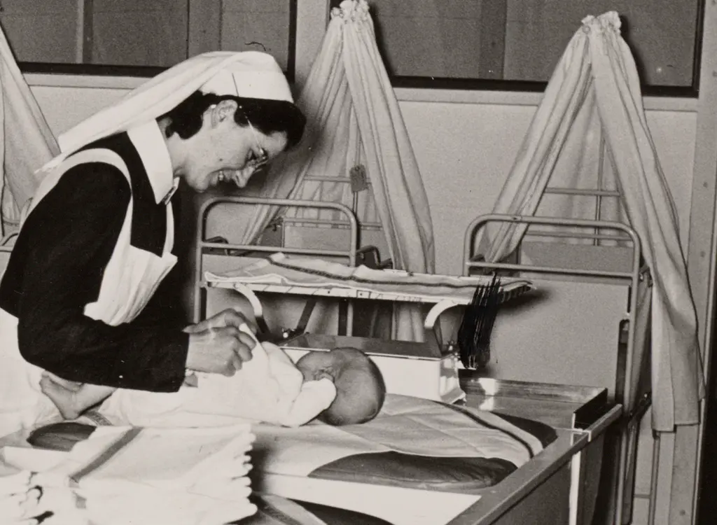 Verpleegster is bezig met kleertjes van een baby die op een tafel ligt met op de achtergrond een aantal wiegbedjes