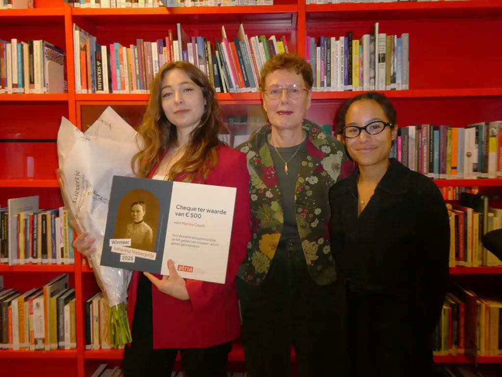 Marlies Couch met bloemen en grote cheque ter waarde van 500 euro, Francisca de Haan, en Alex Erica Mary McQueen, staand voor rode boekenkasten